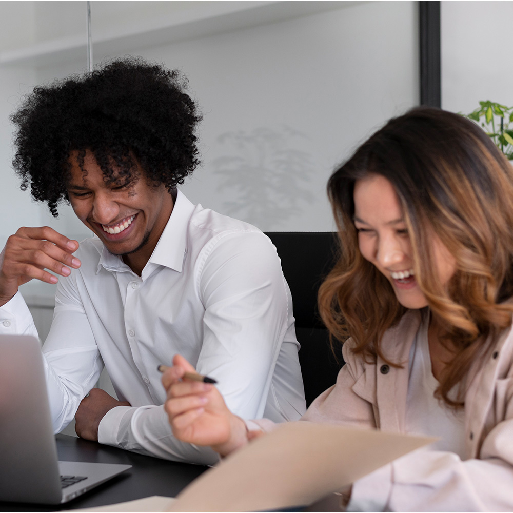 A man and a woman in an office smiling while looking at the same computer screen, collaborating and sharing ideas